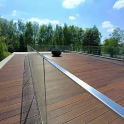 Rozległy taras z desek w ciepłym odcieniu brązu, otoczony szklaną balustradą z metalowymi elementami, na tle zieleni drzew i błękitnego nieba z delikatnymi obłokami. Na środku tarasu stoi czarna...