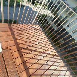 CleanWood - Widok z góry na drewniany taras na balkonie, z metalową balustradą rzucającą cienie na deski, nowoczesna architektura budynków w tle.