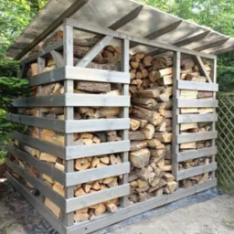 Drewniany regał na opał z przezroczystym zadaszeniem, wypełniony pociętymi kawałkami drewna, otoczony zielenią.