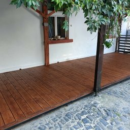 CleanWood - Zadaszony taras z ciemnego drewna z widokiem na kamienną nawierzchnię, porośnięty winoroślą, przylegający do białej ściany budynku z oknem.