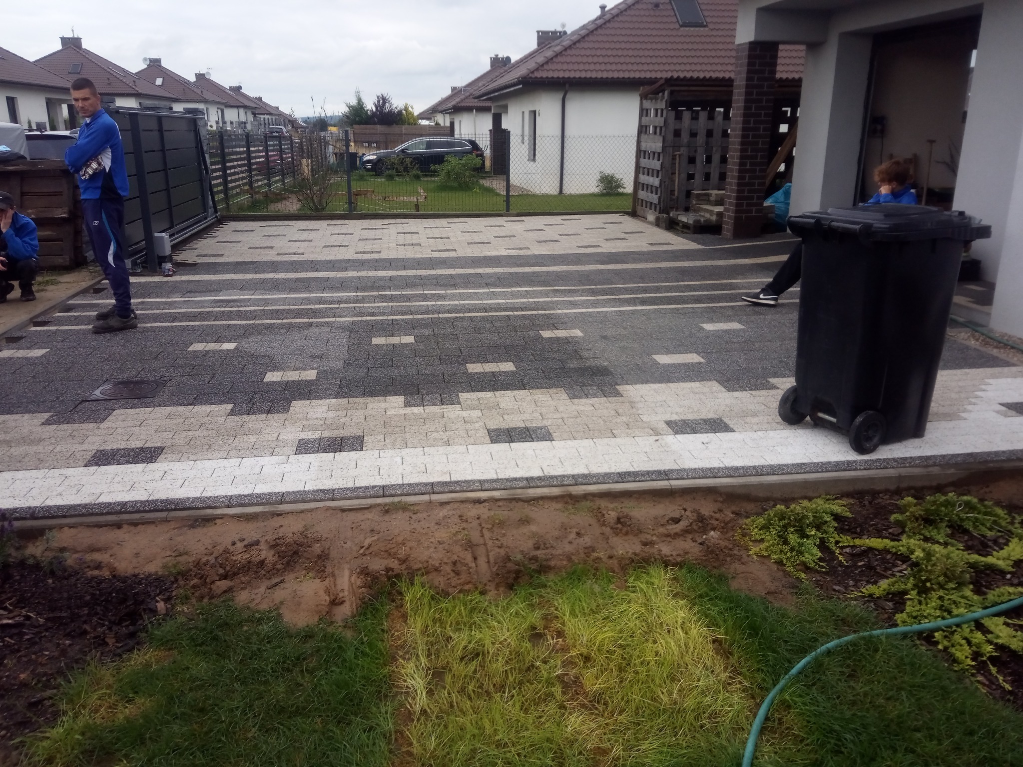 Ułożona kostka brukowa w dwóch odcieniach szarości z widocznymi pracownikami w niebieskich strojach, częściowo wykończony trawnik i domy w tle.