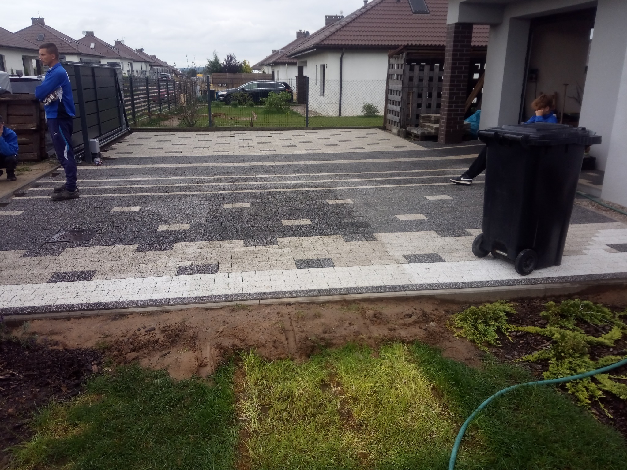 Ułożona kostka brukowa w geometryczny wzór przed domem jednorodzinnym z widoczną przerwą na trawnik i osobą w stroju sportowym po lewej stronie kadru.