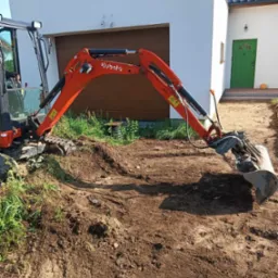 Pomarańczowa minikoparka Kubota podczas prac ziemnych na działce przed domem z zielonymi drzwiami i brązową bramą garażową.