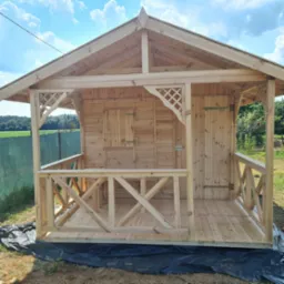 Surowa, drewniana konstrukcja altany z kratownicowymi zdobieniami, widoczne zawiasy i zamknięcia, na tle zieleni i błękitnego nieba.