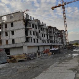 MARIAN PARZYGNAT MP-BUD FIRMA REMONTOWO-BUDOWLANA - Wznoszenie wielopiętrowego budynku z białego bloczka, widoczny żuraw budowlany, teren budowy z materiałami i pojazdem dostawczym. Ujęcie z dołu, pochmurne niebo.