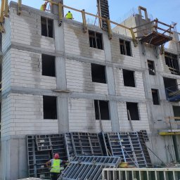 MARIAN PARZYGNAT MP-BUD FIRMA REMONTOWO-BUDOWLANA - Wznoszenie wielopiętrowego budynku z betonu komórkowego. Widoczne rusztowania, szalunki i pracownicy w odblaskowych kamizelkach. Budowa w toku, jasne niebo w tle.