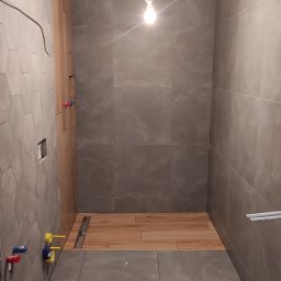 MARIAN PARZYGNAT MP-BUD FIRMA REMONTOWO-BUDOWLANA - Wnętrze łazienki w trakcie wykańczania: szare płytki na ścianach, drewnopodobna podłoga w strefie prysznica, widoczne instalacje wodne i elektryczne. Surowy stan, brak armatury.