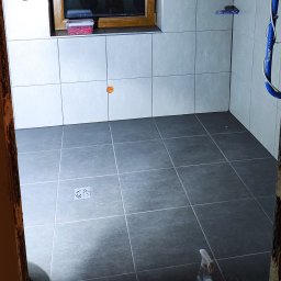 MARIAN PARZYGNAT MP-BUD FIRMA REMONTOWO-BUDOWLANA - Wykończona łazienka z szarymi płytkami na ścianach i podłodze. Widoczny odpływ liniowy, okno i fragment instalacji. Użyto poziomych płytek ściennych.