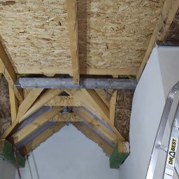 MARIAN PARZYGNAT MP-BUD FIRMA REMONTOWO-BUDOWLANA - Drewniana konstrukcja dachu z elementami metalowymi i płytą OSB, widok z dołu. Fragment ściany z widocznymi śladami szpachlowania i drabina.