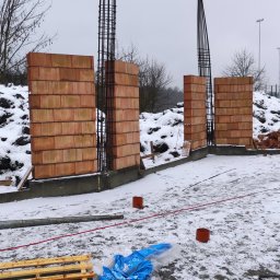 Wznoszenie ścian z czerwonej cegły ceramicznej na fundamencie w zimowej scenerii. Widoczne zbrojenia i zasypany śniegiem teren budowy. Prace murarskie w toku.