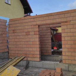Wznoszenie ścian z czerwonej cegły ceramicznej na fundamencie. Widoczny otwór drzwiowy, ocieplenie styropianem i klimatyzator na elewacji budynku. Częściowo ułożone cegły.