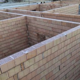 Wznoszenie ścian domu z pustaków ceramicznych w zimowej scenerii. Widoczne fragmenty ścian i otwory okienne. Świeży śnieg na murach. Ujęcie z góry.