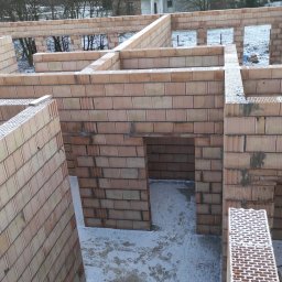 Wnętrze domu w trakcie budowy z cegieł ceramicznych. Widoczne ściany nośne i działowe, otwory okienne i drzwiowe. Na podłodze resztki śniegu. Surowy stan.