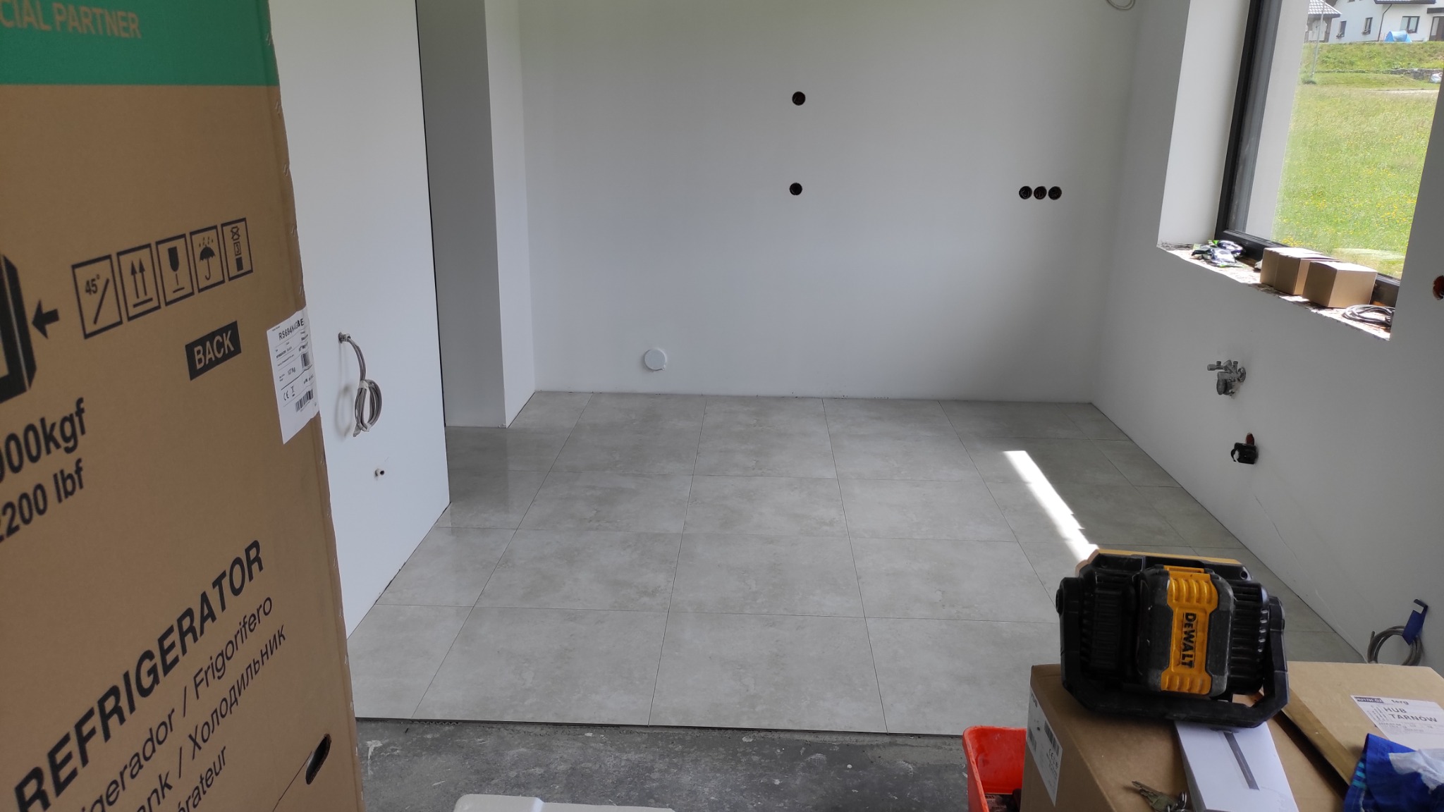 Wykończona podłoga z płytek w jasnym pomieszczeniu z białymi ścianami. Widoczne przygotowane instalacje, okno z widokiem na zewnątrz i karton z napisem REFRIGERATOR.
