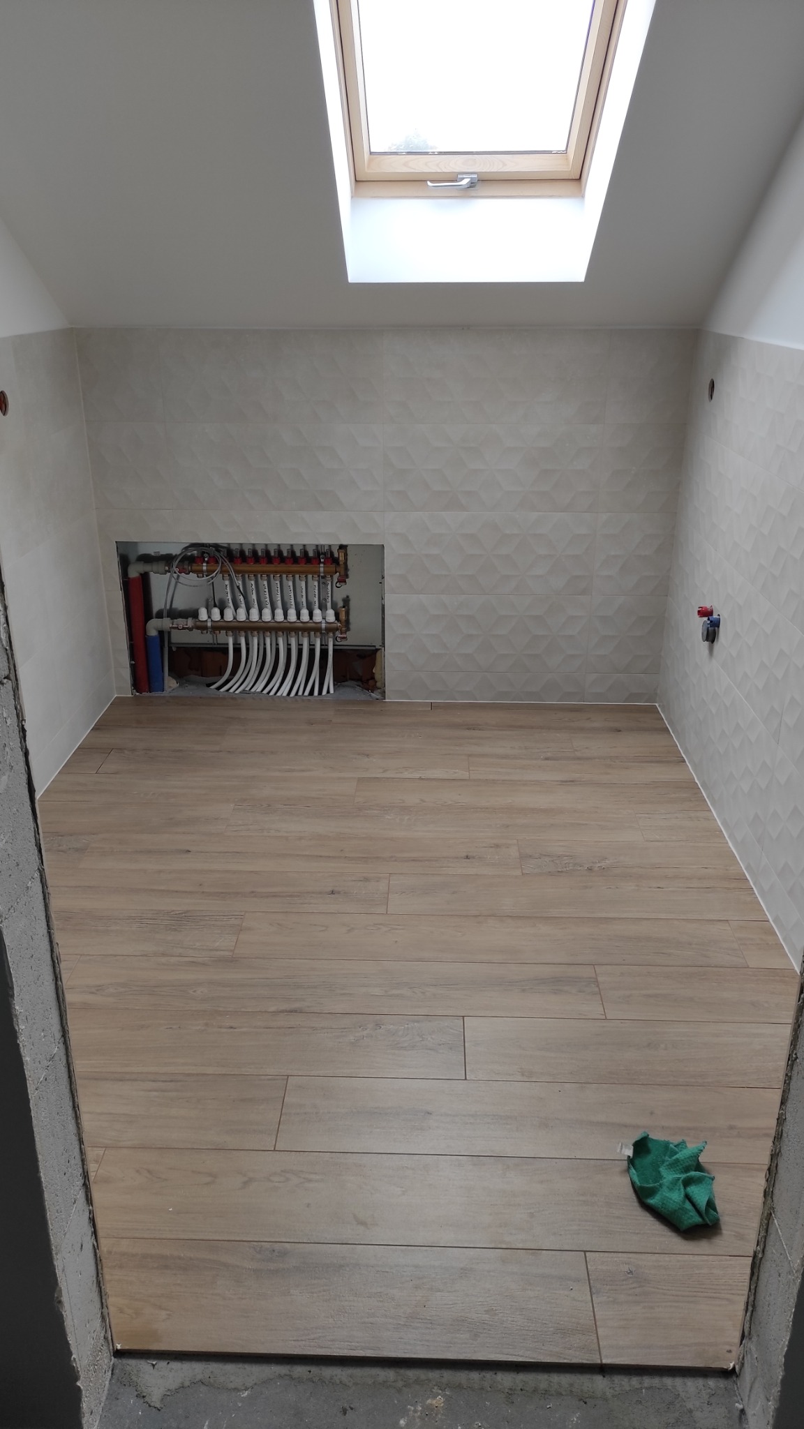 Wykończona łazienka na poddaszu z drewnopodobną podłogą, geometrycznymi płytkami ściennymi i instalacją ogrzewania podłogowego w ścianie. Widoczne okno dachowe.