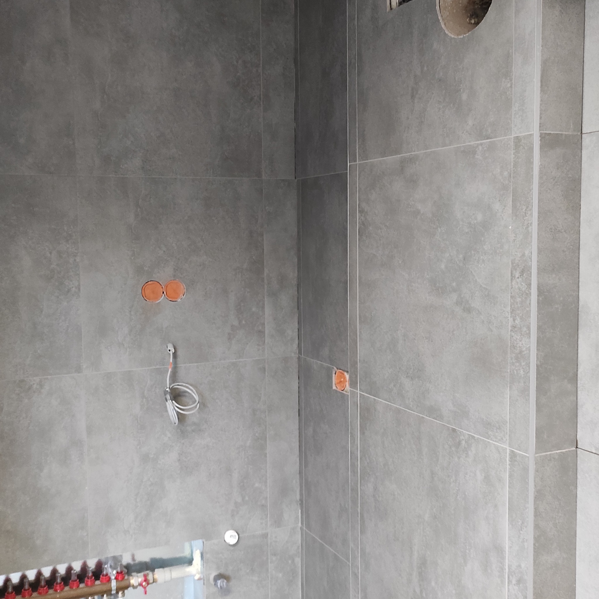 Szare, matowe płytki w łazience, widoczne instalacje wodne i elektryczne. Minimalistyczny design, surowy klimat. Ujęcie z dołu, widoczny fragment rozdzielacza ogrzewania podłogowego.