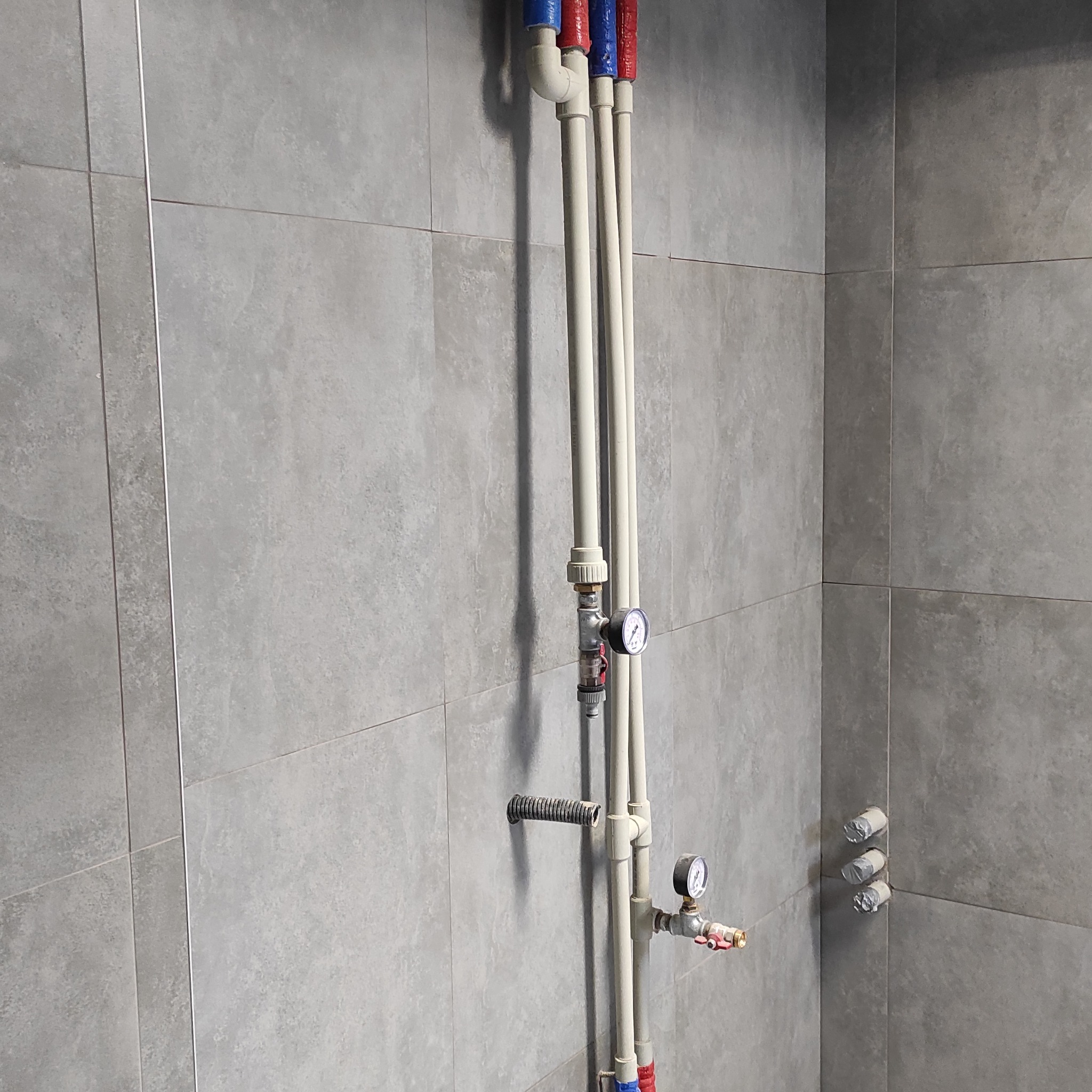 Szczegółowe ujęcie instalacji wodnej z rurami PEX i manometrami na tle szarych, kwadratowych płytek w łazience. Widoczne przyłącza i zabezpieczenia.