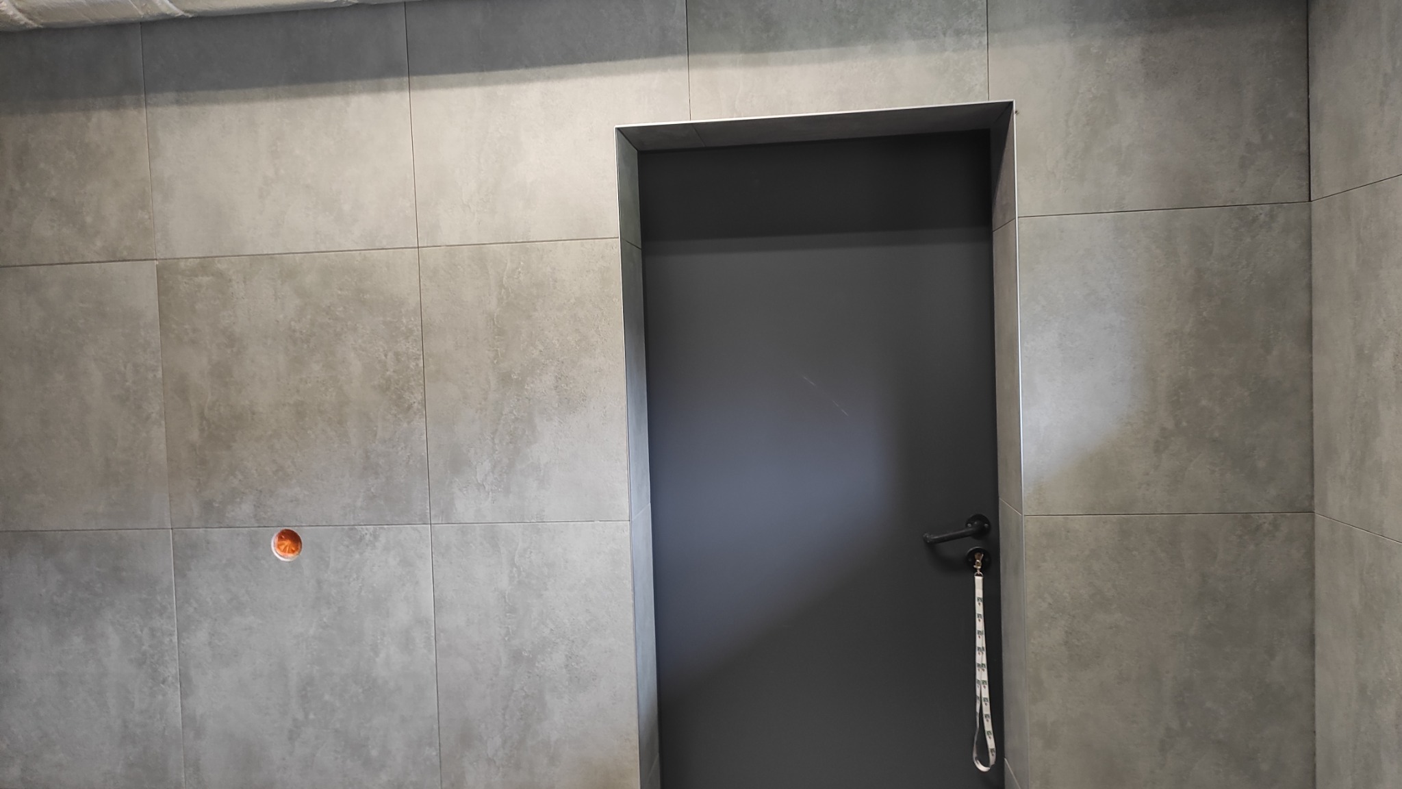 Szare, matowe płytki ścienne w łazience z widocznym otworem na gniazdko i szarą, nowoczesną ościeżnicą z drzwiami i klamką. Minimalistyczny design.