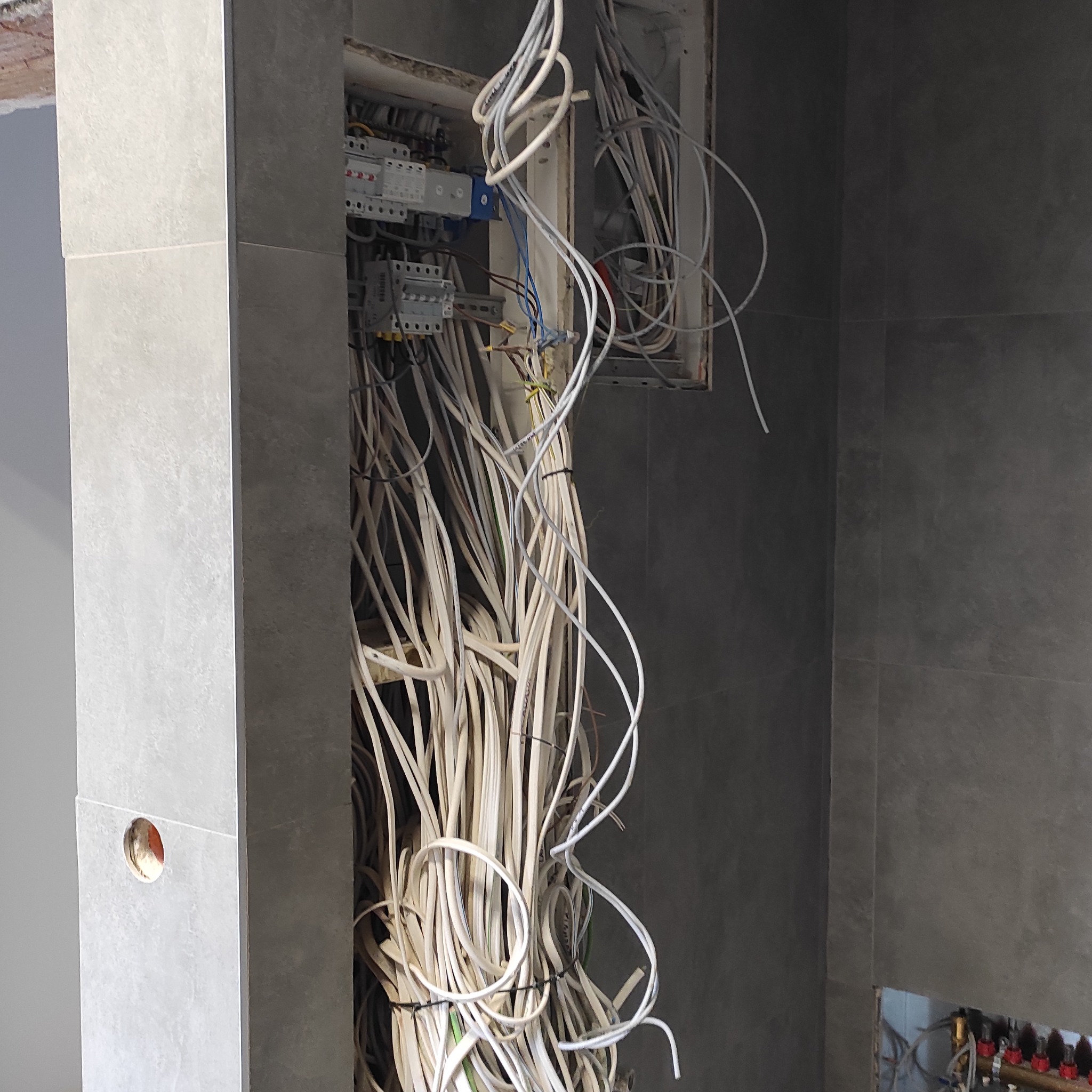 Rozdzielnia elektryczna w trakcie montażu, widoczne liczne przewody i zabezpieczenia. Szare płytki na ścianach. Instalacja w trakcie prac wykończeniowych.