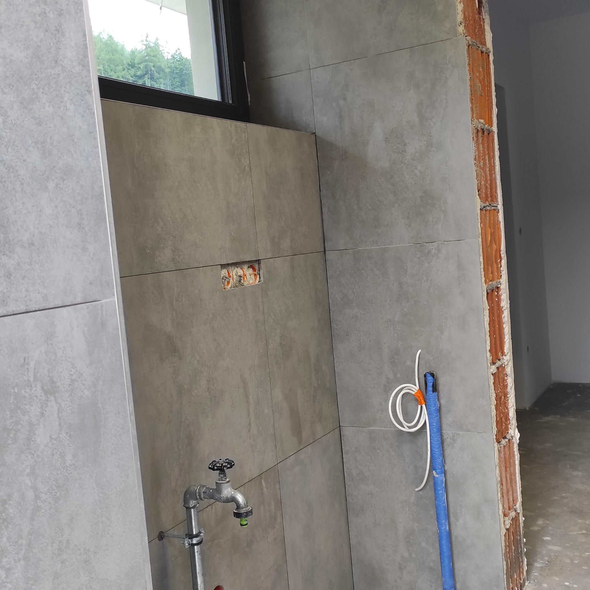 Wykończona ściana z szarych płytek w łazience, widoczny kran, instalacja elektryczna i fragment ściany z cegły. Prace wykończeniowe w nowym domu.