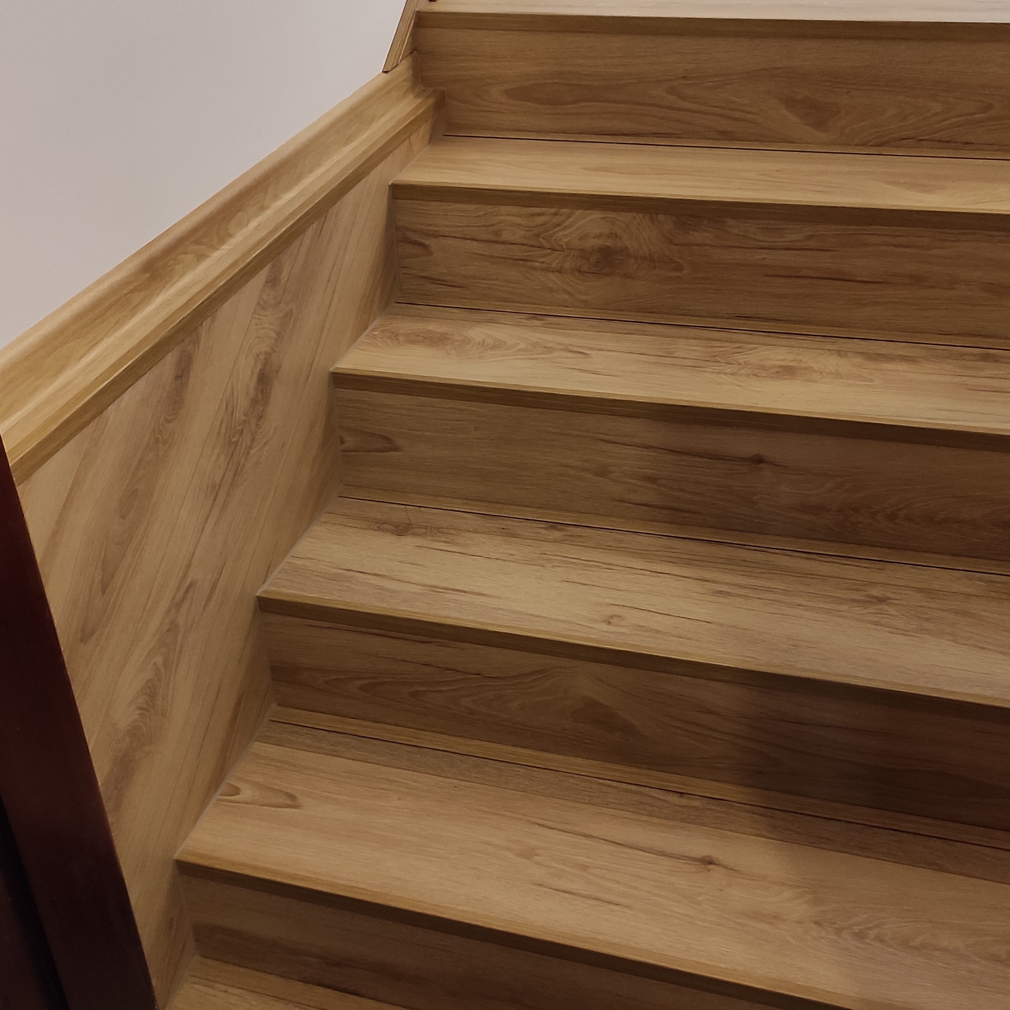 Wykończone schody z drewnopodobnymi panelami, widok z dołu. Estetyczne połączenie paneli na stopniach i pionowych elementach. Jasne, ciepłe odcienie drewna.