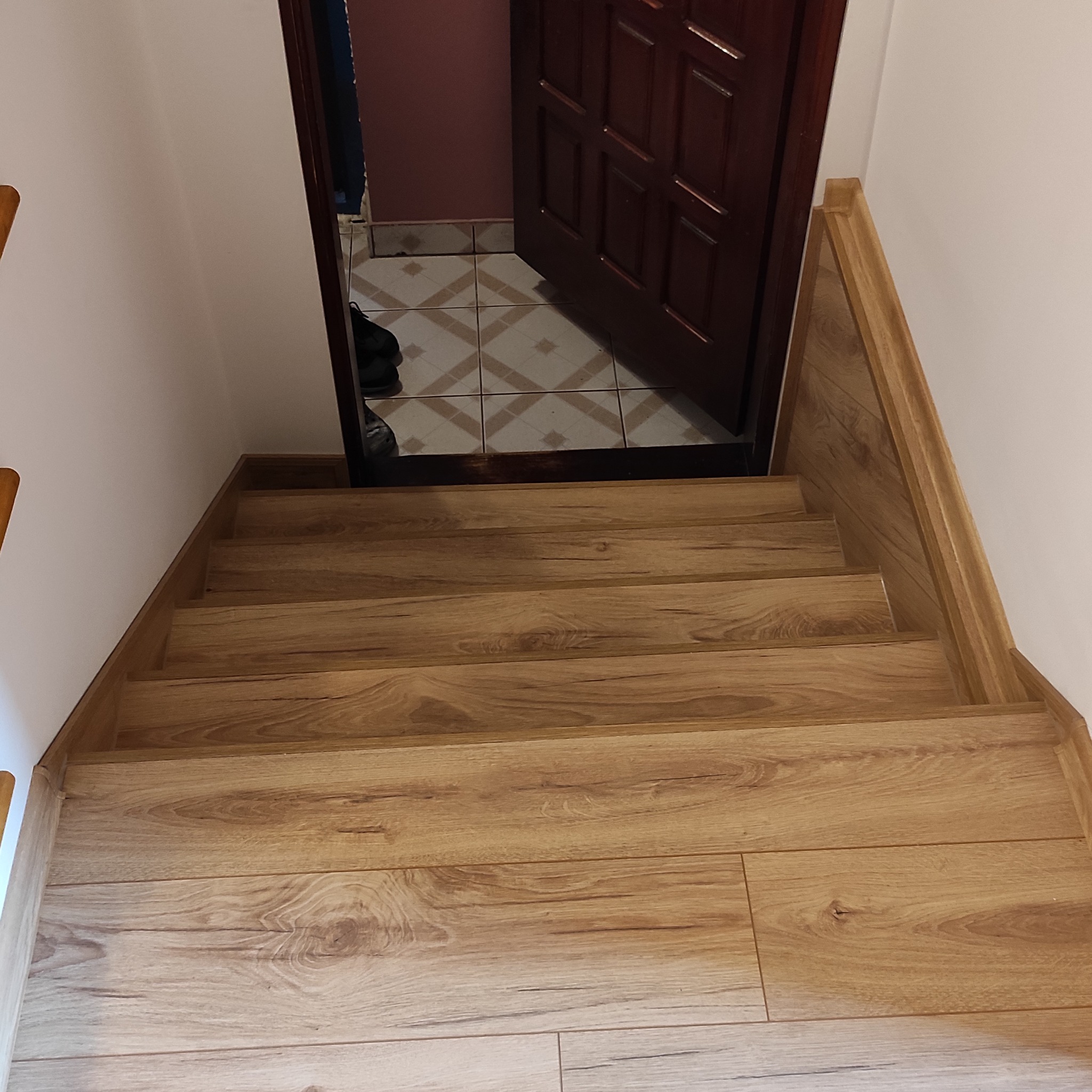 Schody wyłożone panelami w odcieniach drewna, prowadzące w górę, z widocznym fragmentem otwartych drzwi i podłogi w pomieszczeniu na górze. Jasne ściany.