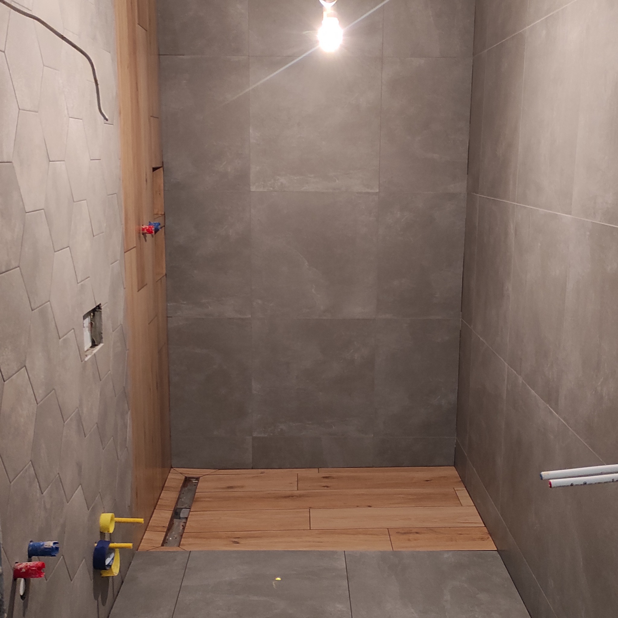 Wnętrze łazienki w trakcie wykańczania: szare płytki na ścianach, drewnopodobna podłoga w strefie prysznica, widoczne instalacje wodne i elektryczne. Surowy stan, brak armatury.