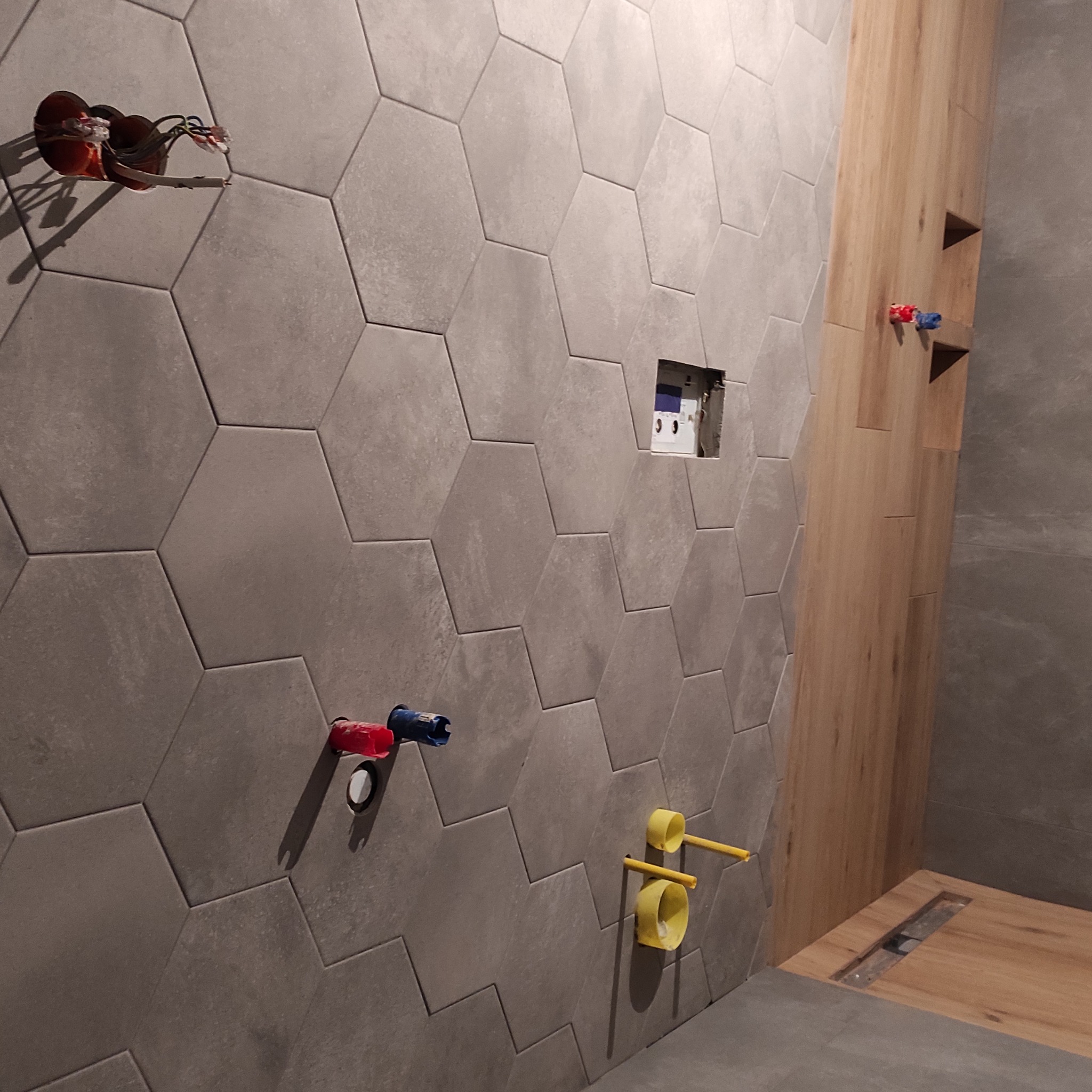 Ściana wyłożona szarymi, heksagonalnymi płytkami z widocznymi instalacjami wodnymi i elektrycznymi. Fragment drewnianej zabudowy z wnękami i odpływem liniowym.