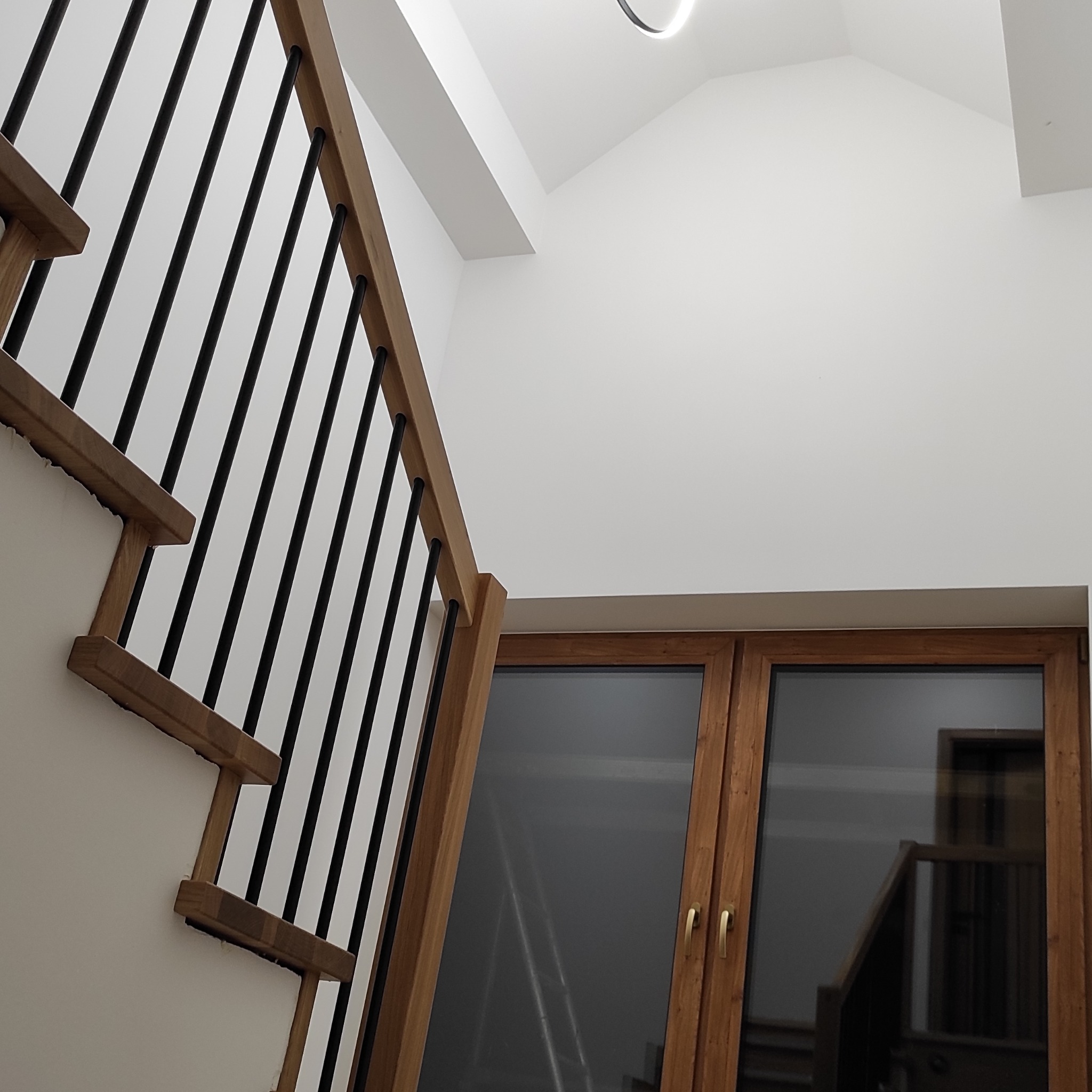 Nowoczesne schody z drewnianymi stopniami i czarnymi balustradami, widoczne z dołu, z oknem w tle. Minimalistyczny design i jasne wnętrze.