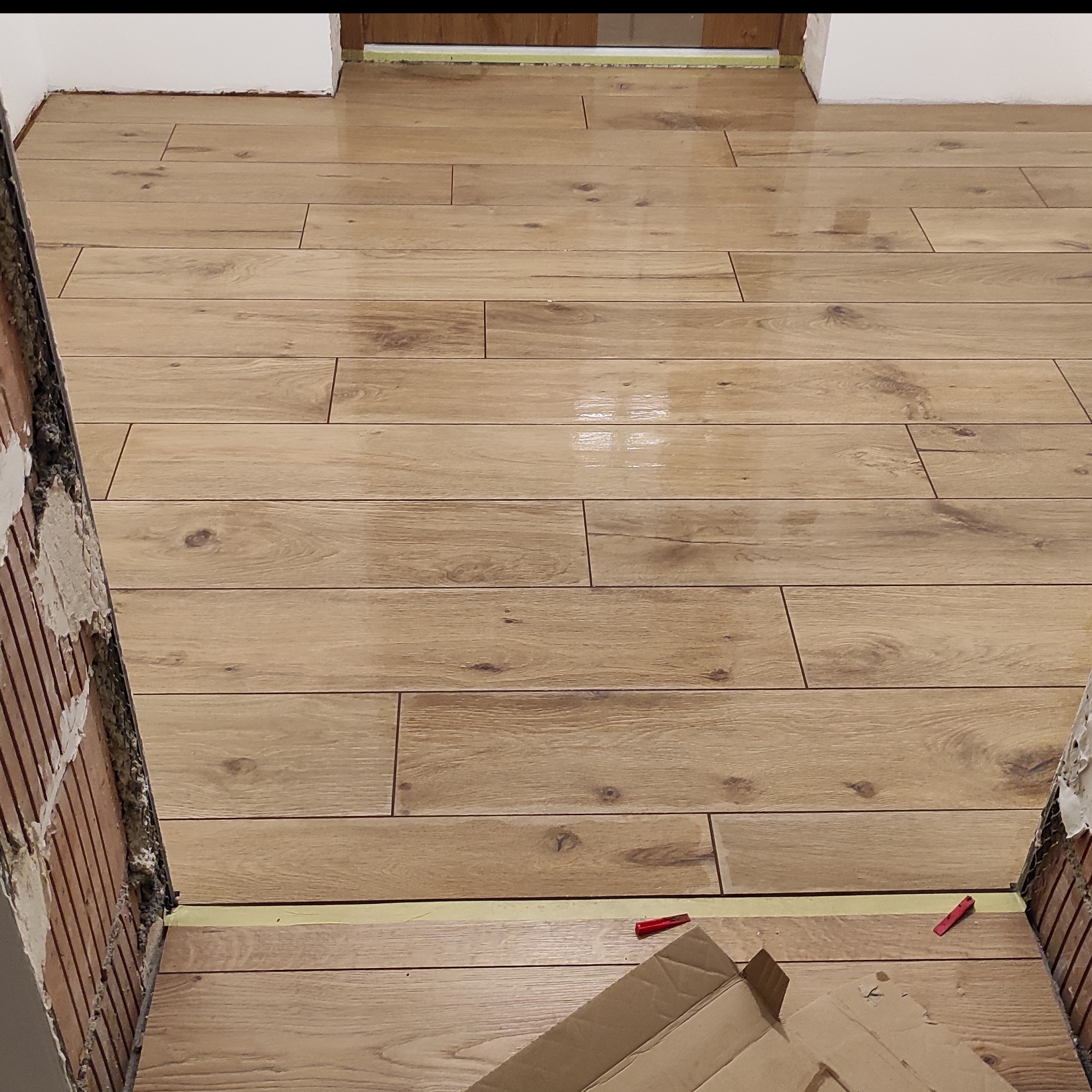 Ułożona podłoga z płytek imitujących drewno, widok z progu. Fragment ściany z widocznym klejem. Na podłodze karton i czerwone kliny dystansowe.