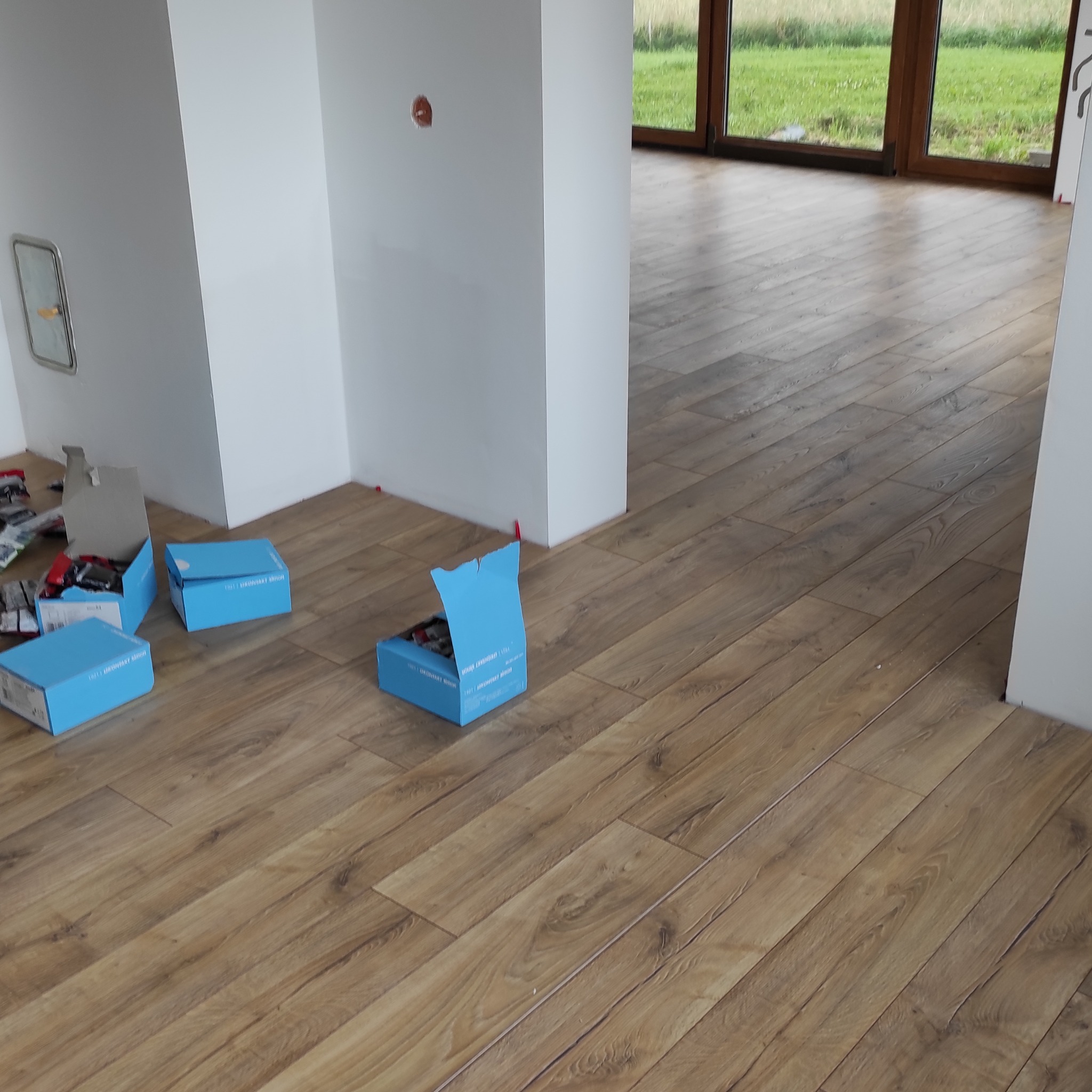 Układanie paneli podłogowych w Skawie. Widoczne otwarte pudełka z panelami i akcesoriami. Podłoga w odcieniach brązu, imitująca drewno, ułożona w jodełkę.