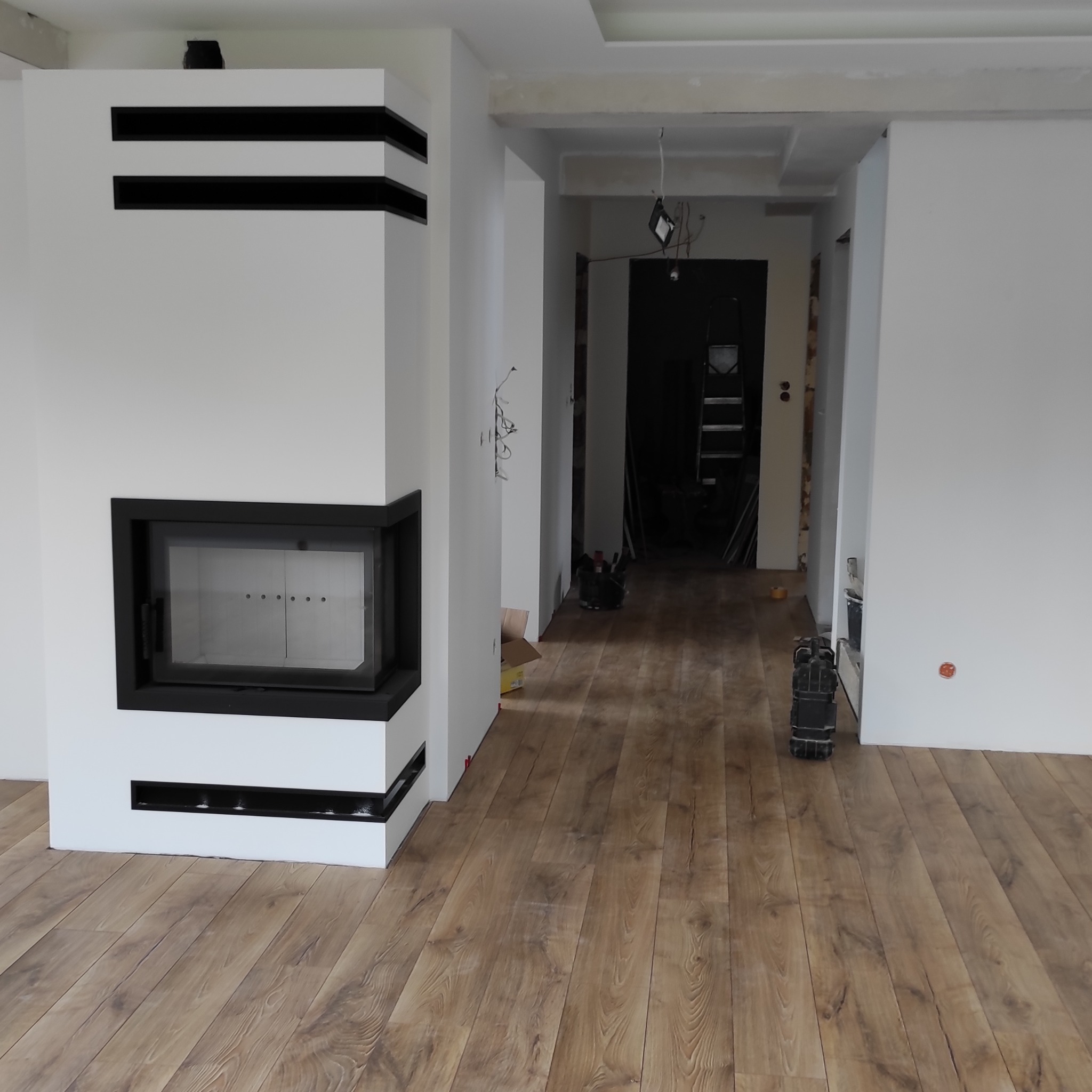 Nowoczesny kominek z czarnymi akcentami w jasnym wnętrzu z panelami podłogowymi. Widoczne ślady prac wykończeniowych.