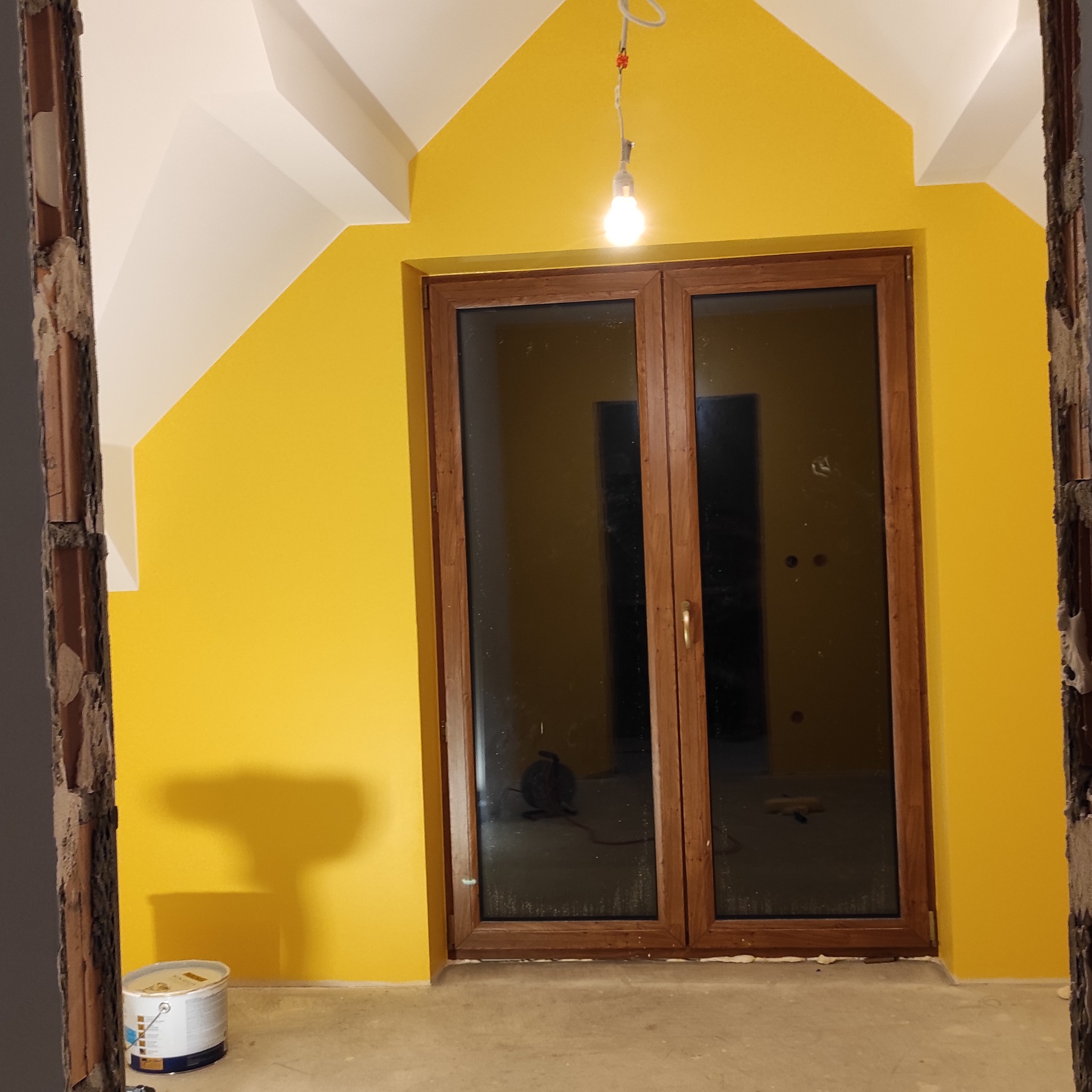 Żółta ściana po malowaniu z drewnianymi drzwiami balkonowymi, widok przez otwór w ścianie. Na podłodze puszka farby. W tle widoczny przedłużacz.
