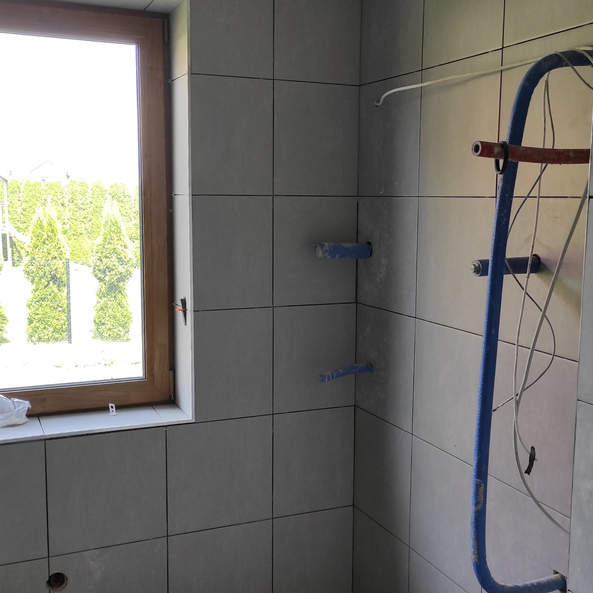 Ściana w łazience wyłożona szarymi płytkami, widoczne wystające rury instalacji wodnej i okno z widokiem na zieleń. Prace wykończeniowe w trakcie realizacji.