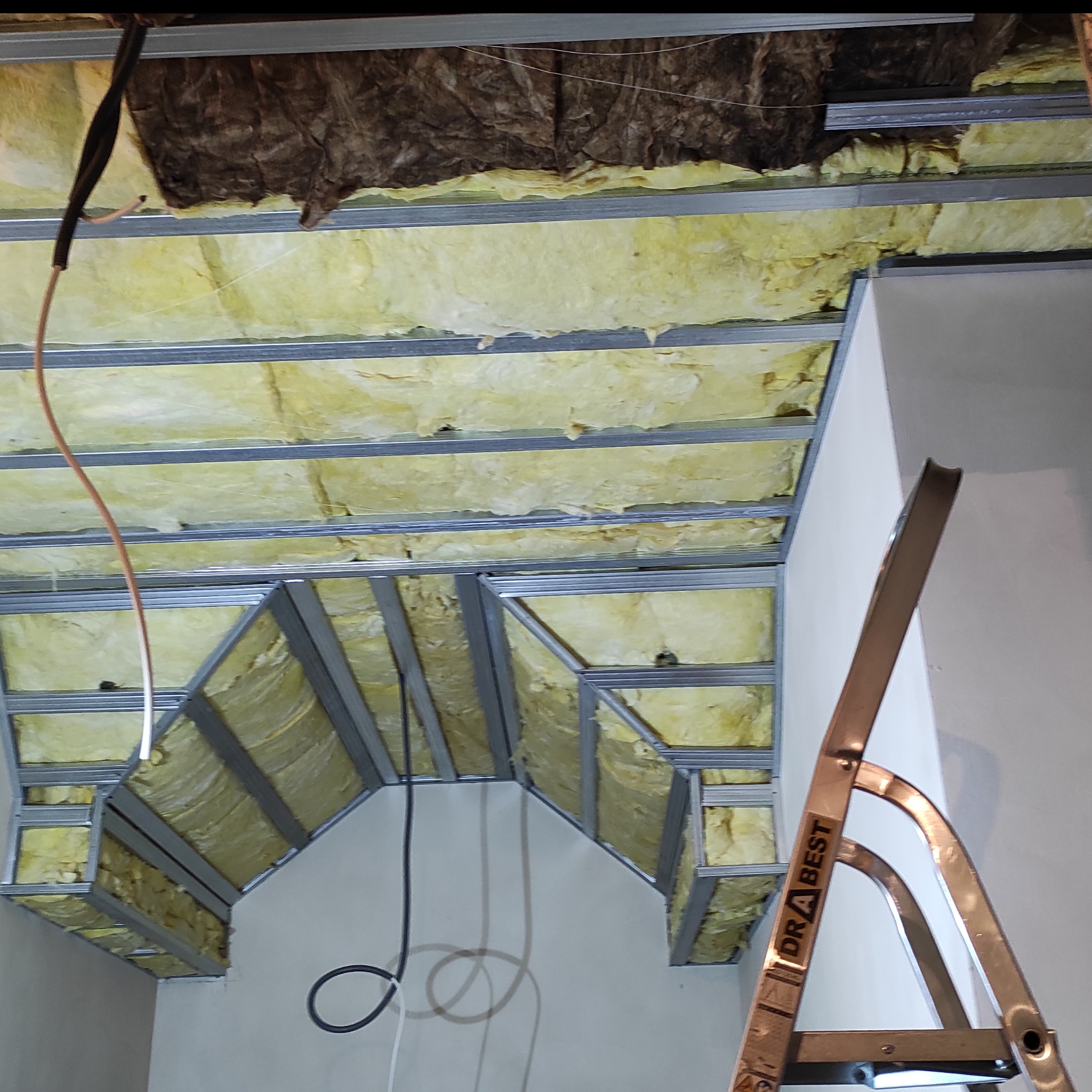 Sufit podwieszany z izolacją z wełny mineralnej, widoczne metalowe profile i fragment drabiny. Prace wykończeniowe w trakcie realizacji.