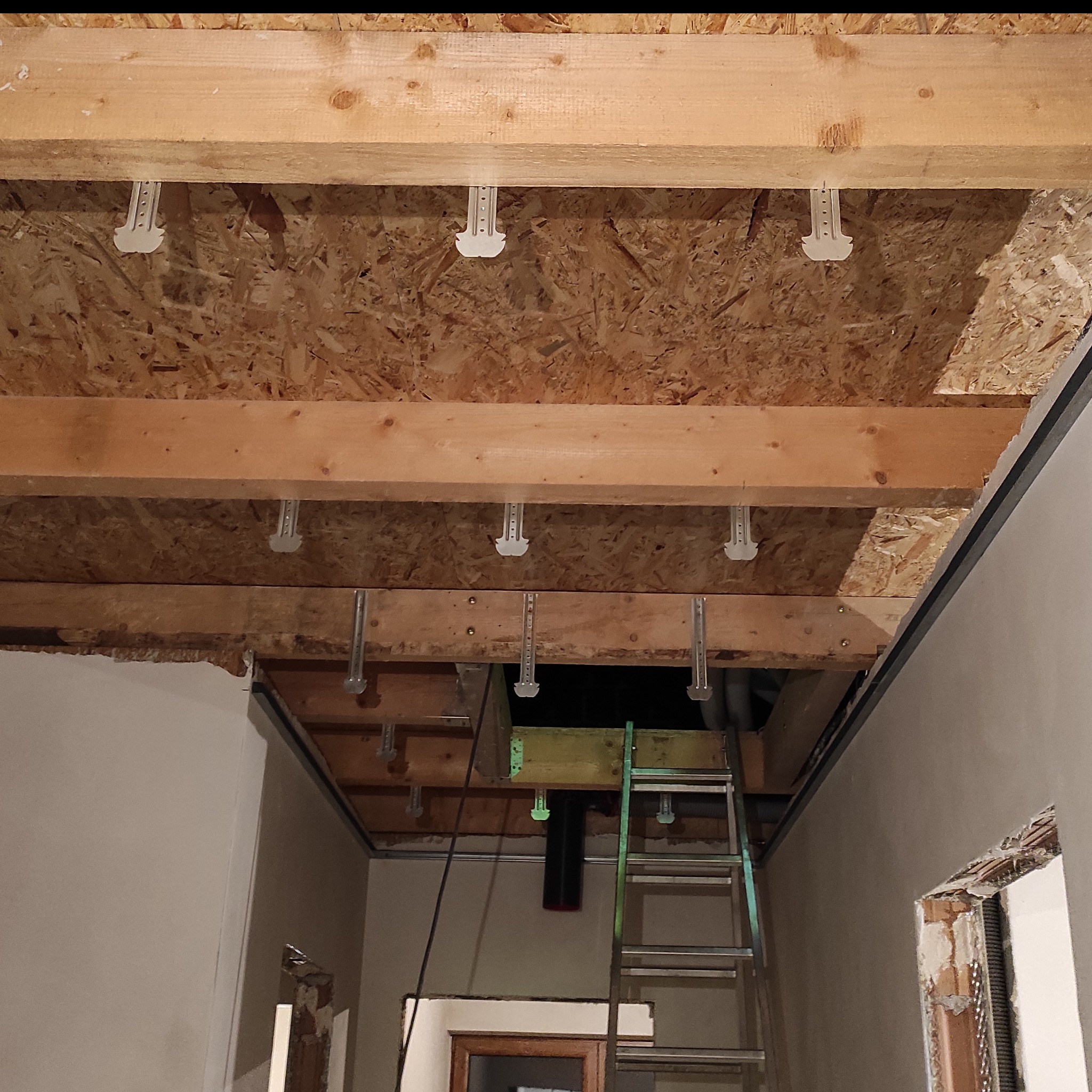 Drewniana konstrukcja sufitu podwieszanego z widocznymi elementami mocującymi, drabina w tle. Prace remontowe w trakcie realizacji, surowy stan wykończenia.