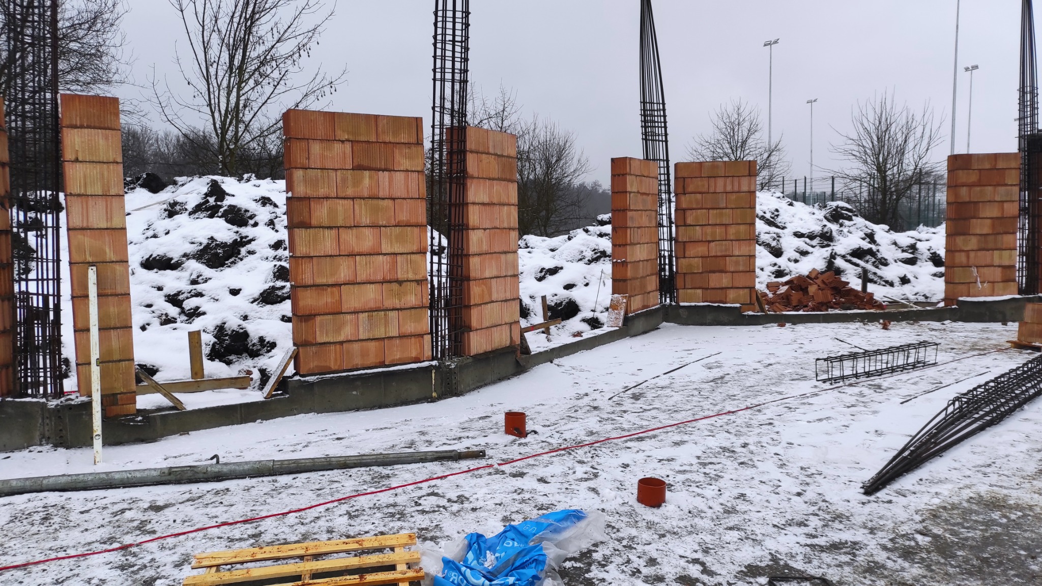 Wznoszenie ścian z czerwonej cegły ceramicznej na fundamencie w zimowej scenerii. Widoczne zbrojenia i zasypany śniegiem teren budowy. Prace murarskie w toku.