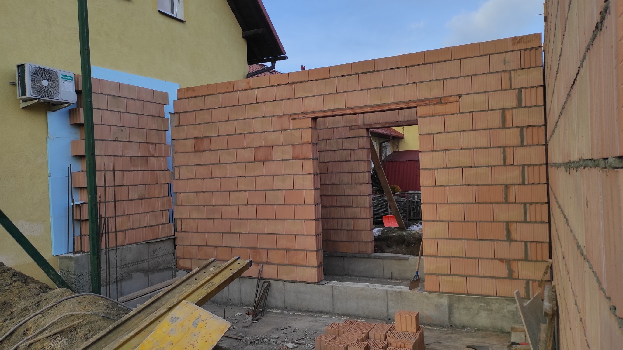 Wznoszenie ścian z czerwonej cegły ceramicznej na fundamencie. Widoczny otwór drzwiowy, ocieplenie styropianem i klimatyzator na elewacji budynku. Częściowo ułożone cegły.
