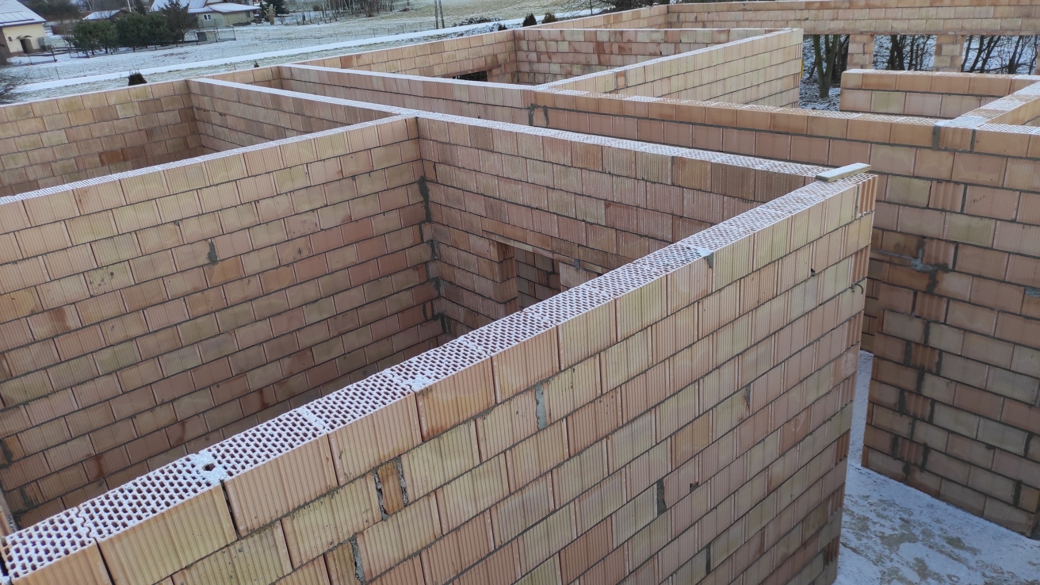 Wznoszenie ścian domu z pustaków ceramicznych w zimowej scenerii. Widoczne fragmenty ścian i otwory okienne. Świeży śnieg na murach. Ujęcie z góry.