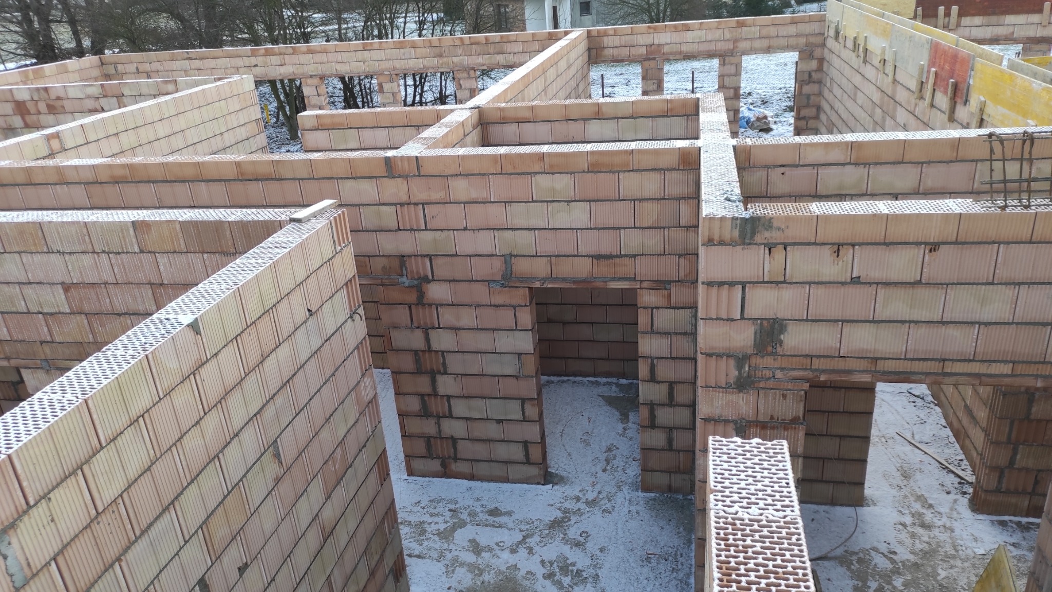 Wnętrze domu w trakcie budowy z cegieł ceramicznych. Widoczne ściany nośne i działowe, otwory okienne i drzwiowe. Na podłodze resztki śniegu. Surowy stan.