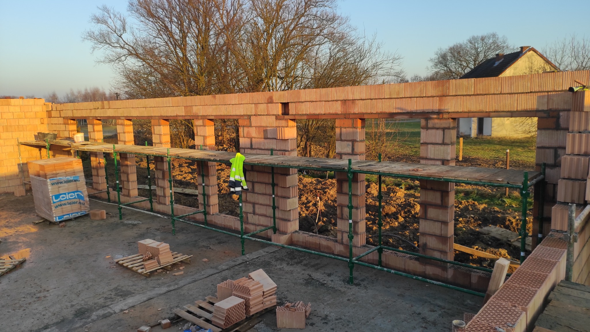 Wznoszenie ścian z czerwonej cegły na budowie domu jednorodzinnego. Widoczne rusztowania, palety z cegłami i fragment krajobrazu w tle. Ujęcie z góry, ciepłe światło.