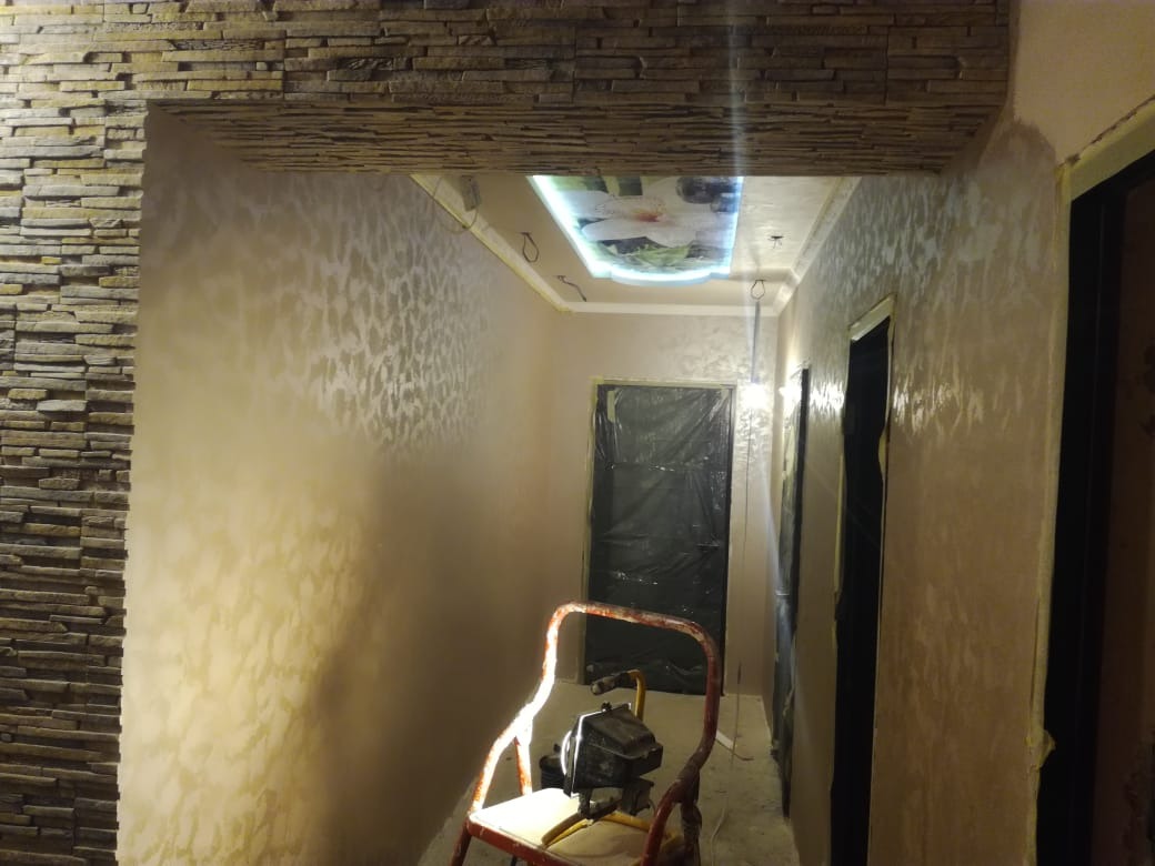 Korytarz w Czarnym Dunajcu po nałożeniu dekoracyjnej gładzi z efektem cieniowania, widoczna ściana wykończona kamieniem dekoracyjnym oraz sprzęt do natryskowego nakładania gładzi.