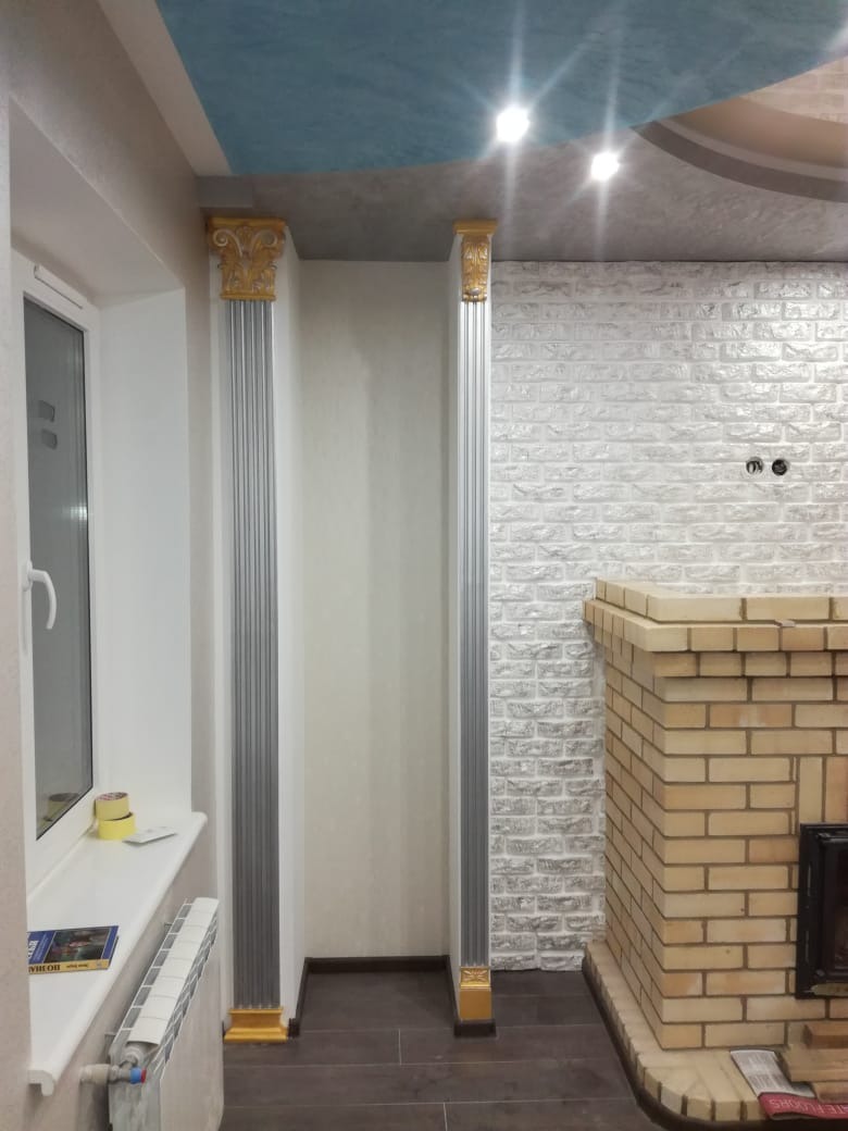 Wykończenie wnętrza z kolumnami dekoracyjnymi w kolorze srebrnym i złotym, ceglana ściana pomalowana na biało oraz sufit z efektem malarskim w odcieniach błękitu i szarości.