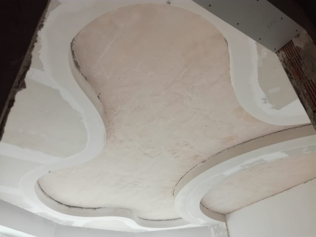 Sufit z dekoracyjnymi elementami z płyty karton gips, w trakcie szpachlowania, widoczne niedoskonałości powierzchni i surowy mur przy krawędzi kadru.