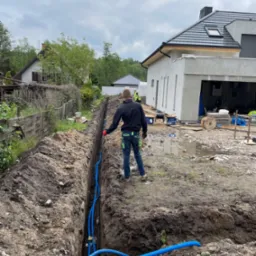 Wykop z niebieskimi rurami kablowymi ułożonymi w ziemi, widok z perspektywy pracownika w odzieży roboczej na tle budowy domu jednorodzinnego