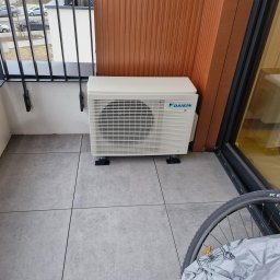 PROKLIM Łukasz Śliwiński - Jednostka zewnętrzna klimatyzacji Daikin na balkonie z szarą podłogą i brązową ścianą. Widoczne barierki balkonu i fragment roweru przy oknie.
