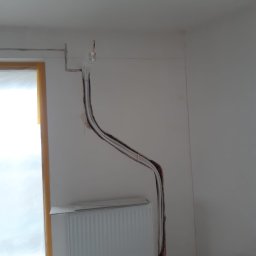 PROKLIM Łukasz Śliwiński - Biały grzejnik ścienny podłączony do instalacji w ścianie, widoczne przewody. Montaż systemu grzewczego w trakcie prac wykończeniowych, wnętrze pomieszczenia.