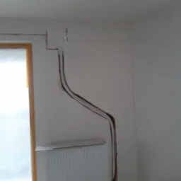 Biały grzejnik ścienny podłączony do instalacji w ścianie, widoczne przewody. Montaż systemu grzewczego w trakcie prac wykończeniowych, wnętrze pomieszczenia.