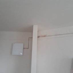 PROKLIM Łukasz Śliwiński - Biały rekuperator ścienny z instalacją, widok z dołu, częściowo widoczna drabina malarska. Ściany w trakcie prac wykończeniowych, widoczne ślady po instalacjach.