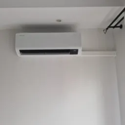 Biała klimatyzacja Samsung WindFree na ścianie z subtelnym wzorem fal, widoczny fragment karnisza i zasłony. Wyświetlacz pokazuje 17 stopni Celsjusza.